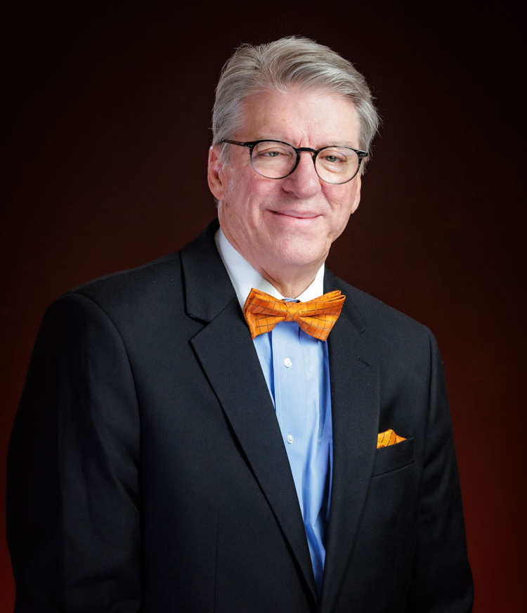 headshot of gray haired gentleman in suit with shadowy orange background behind him.