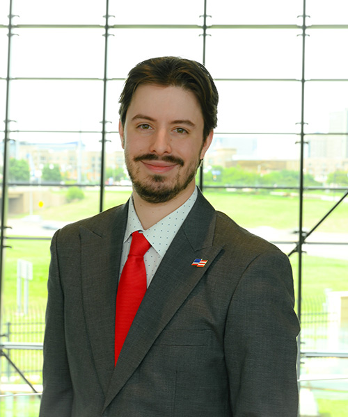 Headshot of younger light skinned man with dark hair standing in front of a large window backdrop
