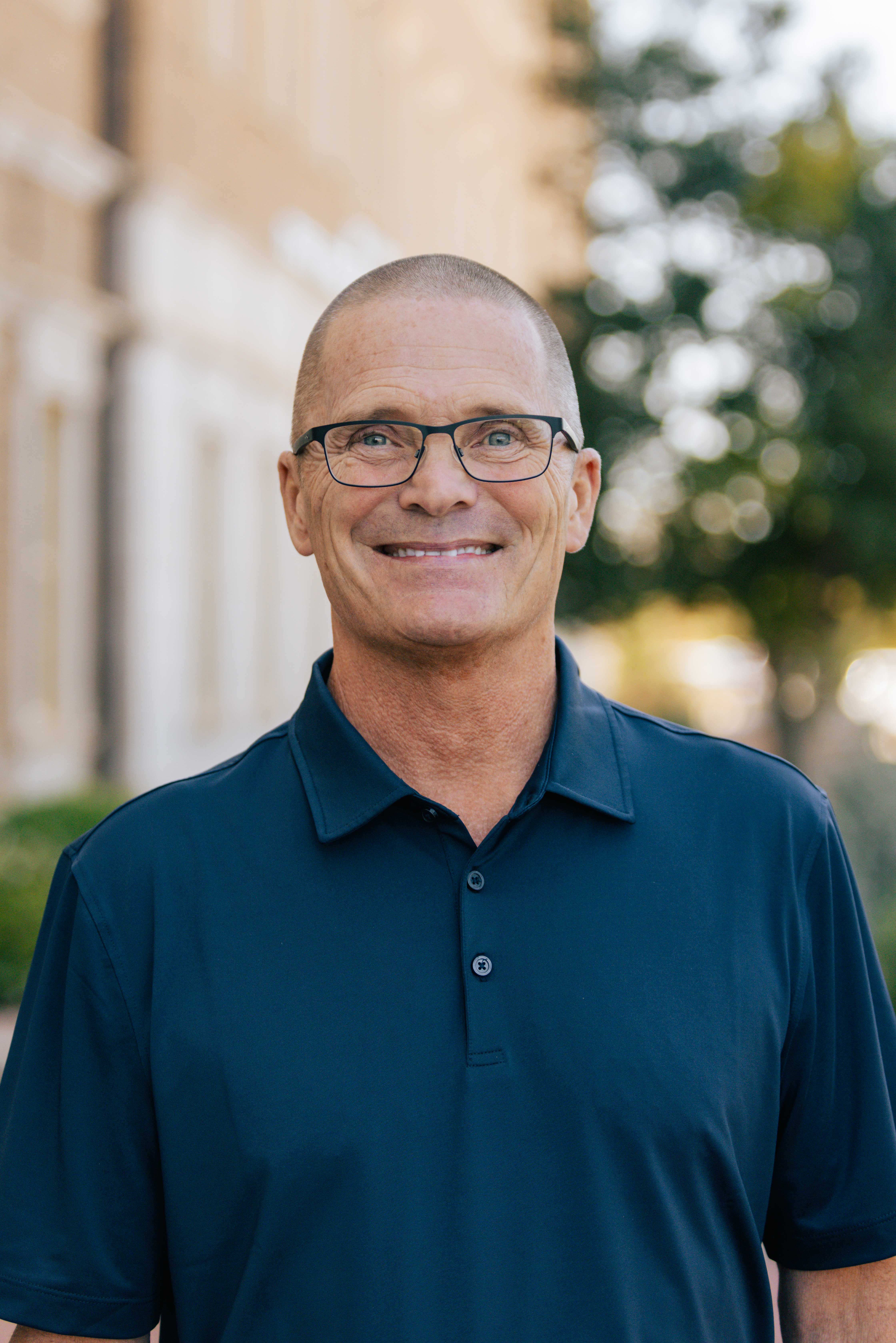 Brad Ewing Outdoor portrait of man with shaved head wearing glasses and blue polo shirt.