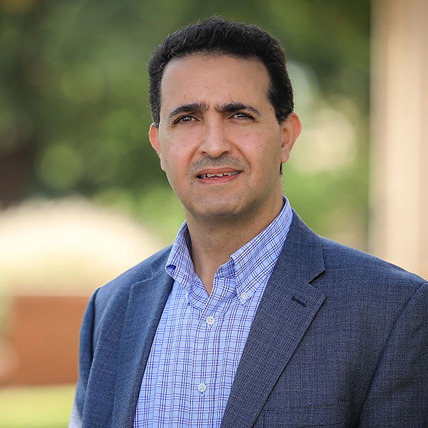 hasan-atiyeh.jpg Headshot of man with black hair wearing blue blazer with blurred foliage in background
